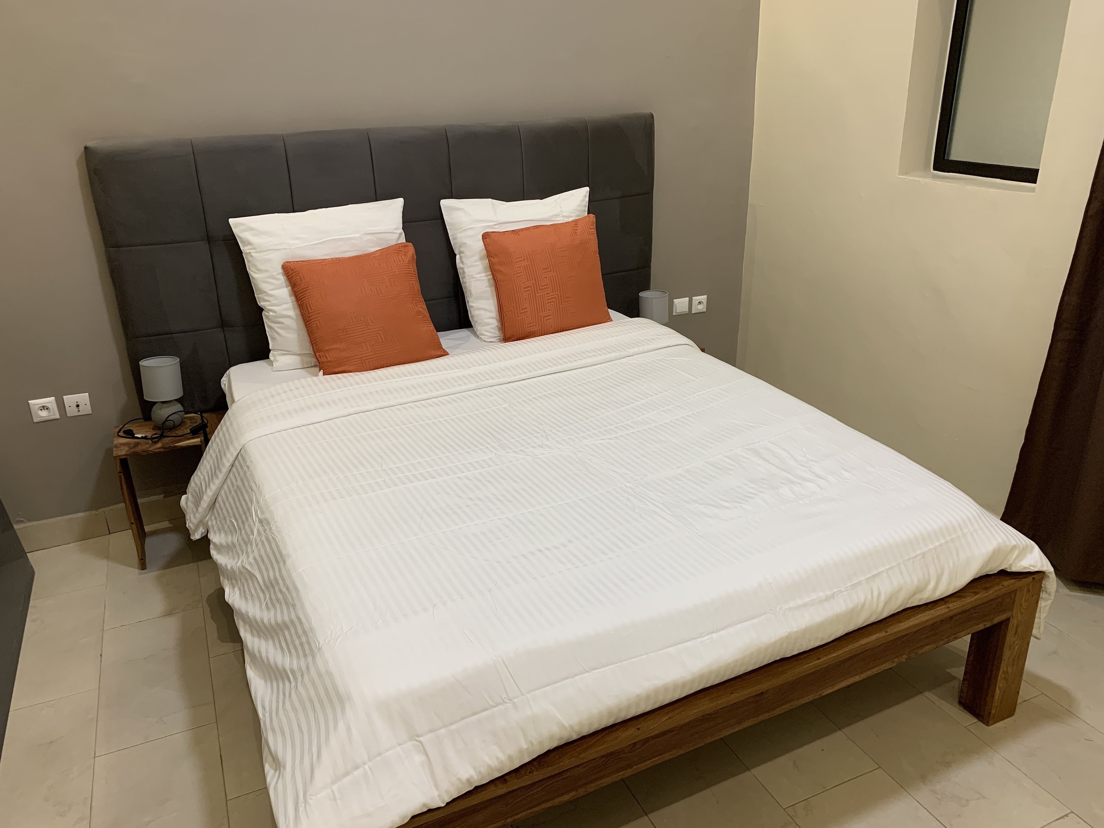 A neatly made bed with a white comforter and two pillows, one white and one orange, in a room with a dark headboard and a small bedside table with a lamp.