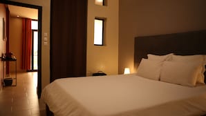 Una habitación de hotel con una cama grande, una mesita de noche, una lámpara y una pequeña mesa con una silla. Hay una puerta que da a otra habitación y una ventana con cortinas.