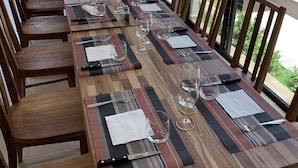 Una larga mesa de comedor con sillas, adornada con copas de vino y cubiertos, en una habitación con ventanas amplias que ofrecen vistas a vegetación y una masa de agua.