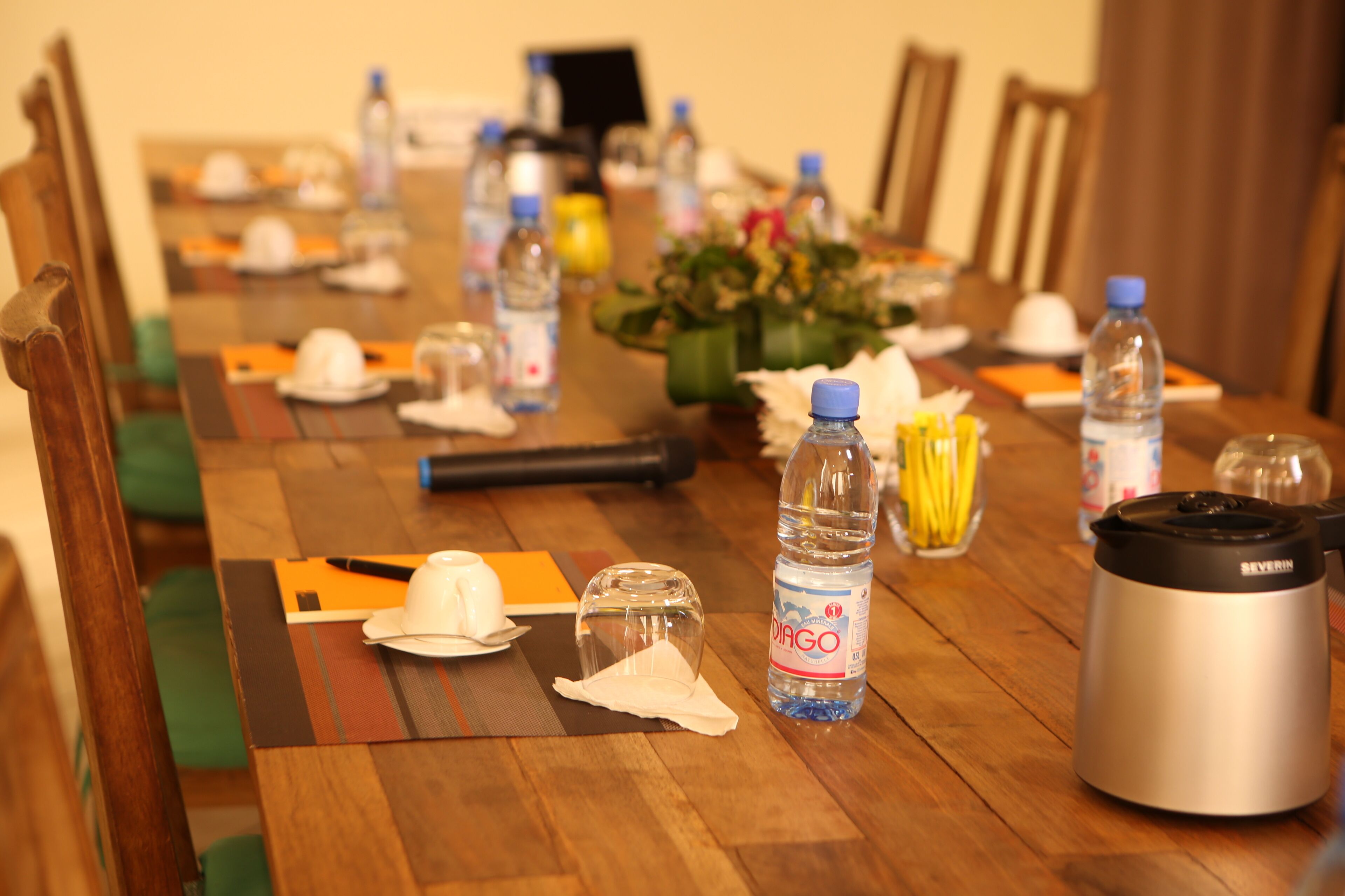 Una mesa de conferencias con botellas de agua, una cafetera y un arreglo floral.