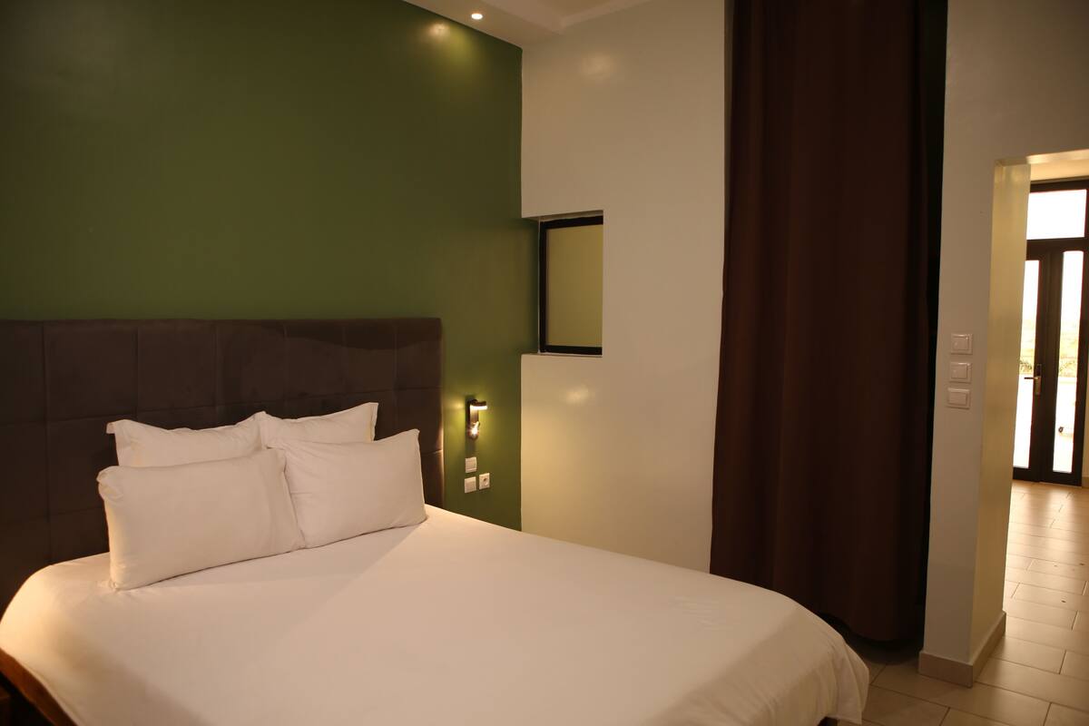 Una habitación de hotel con una cama grande, una pared verde decorativa, una mesita de noche con lámpara y vistas al pasillo.