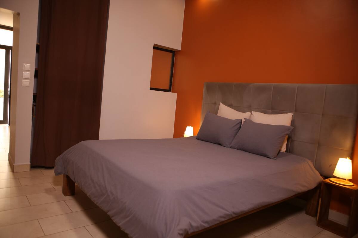 Una cama bien hecha con un cabecero gris y almohadas, una lámpara de noche y un pequeño cuadro en la pared.