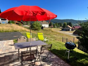 Un patio exterior con una mesa, sillas, un paraguas rojo y una parrilla de barbacoa.