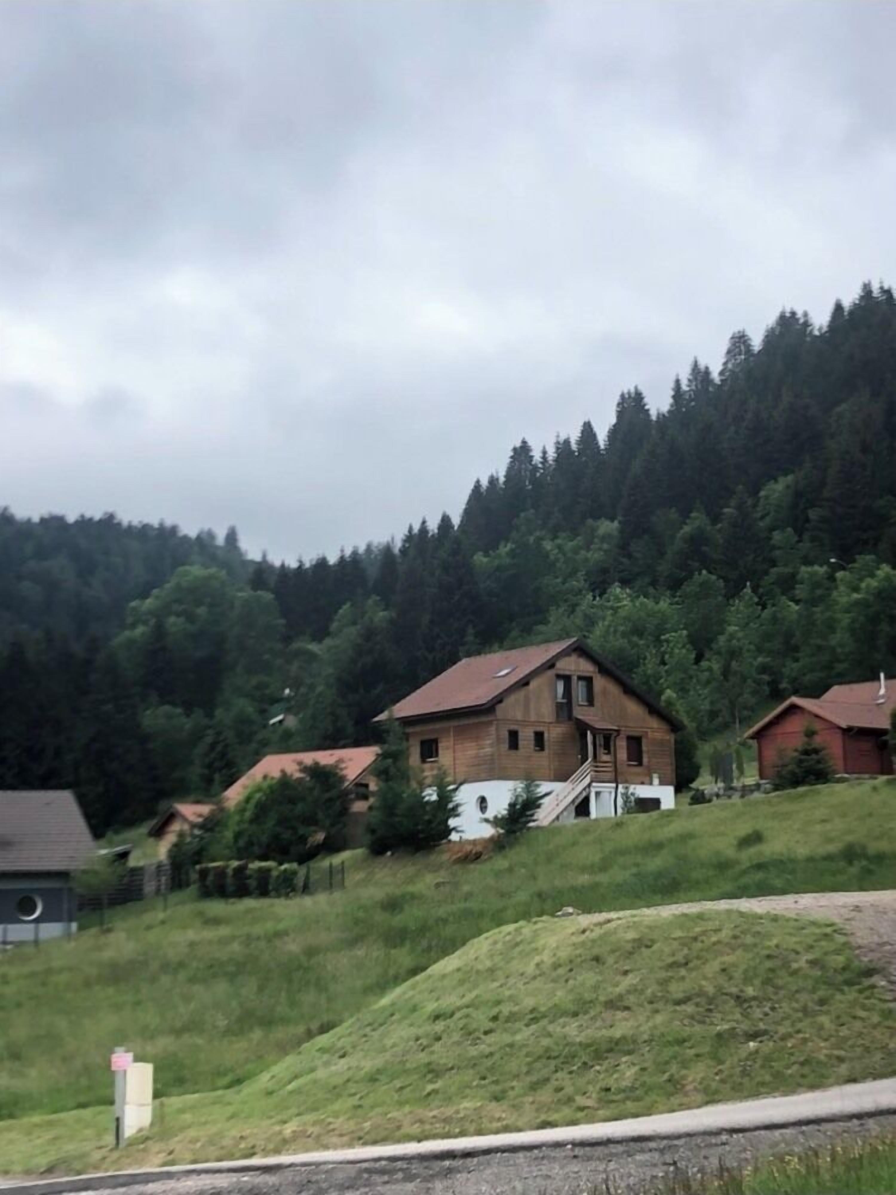 A wooden house with a sloped roof, surrounded by a forested hillside.