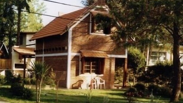 A two-story house with a wooden exterior and a tiled roof, surrounded by greenery and a paved driveway.