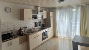 A modern kitchen with white cabinets, a microwave, an oven, and a sink. There is a clock on the wall and a window with curtains. - Susi's Apartment - 60m2 for 2 persons, quiet location and handicapped accessible (Waldgirmes)