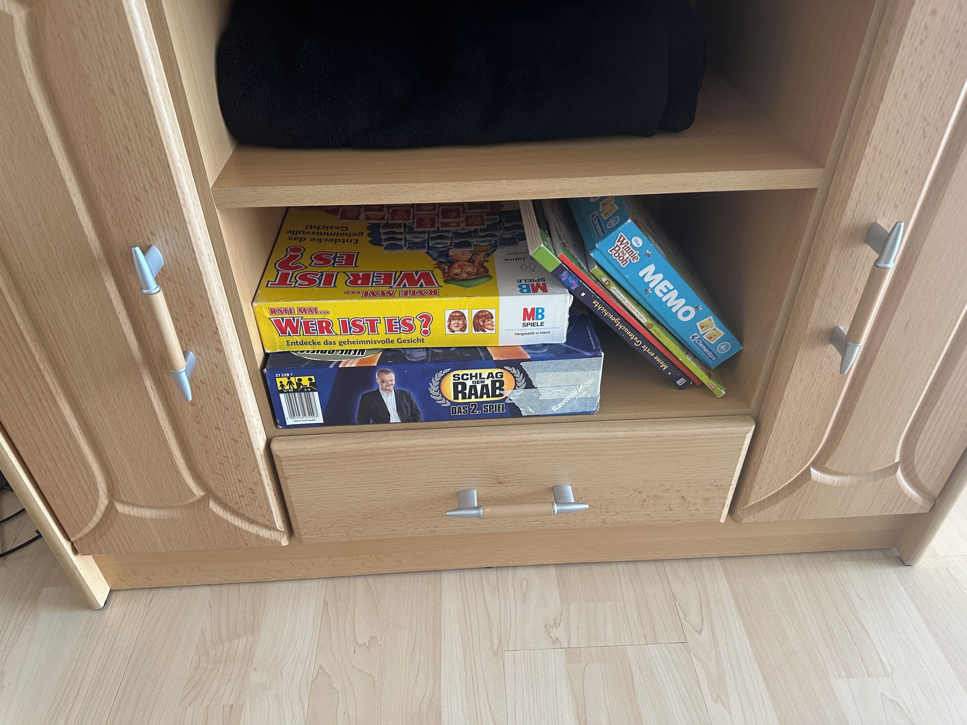 Ein Holzkabinett mit einer Schublade, die verschiedene Gesellschaftsspiele und ein schwarzes Objekt oben darauf enthält.