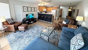 A modern living room with a sofa, ottoman, coffee table, and a TV. There is a kitchen area with a microwave, refrigerator, and bar stools. - Good Night Sleep Tight | Guadalupe Riverfront | WiFi | Pools (New Braunfels)