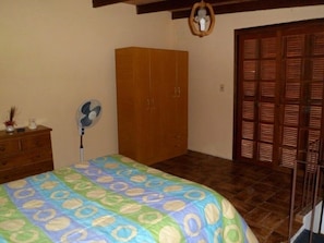 Een slaapkamer met een bed, een houten kast, een ventilator en een deur met luiken.