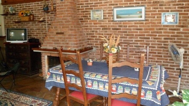 Un coin repas avec un mur en briques, des chaises en bois, une table recouverte d’une nappe à motifs et un téléviseur posé sur un meuble.