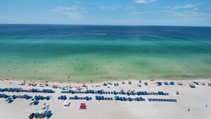 Une plage bordée de rangées de chaises de plage et de parasols bleus, un ciel d’un bleu limpide et une mer calme.