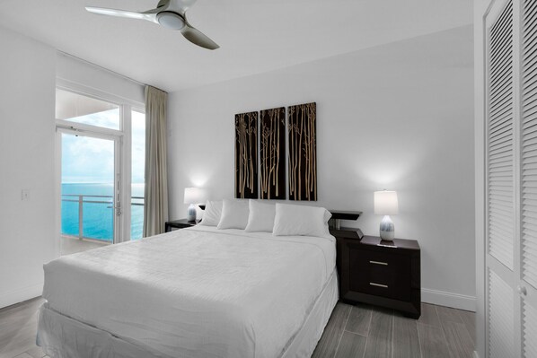 A modern hotel room with a large bed, two bedside tables with lamps, a ceiling fan, and a view of the ocean through a sliding door. - 2211 - Panoramic Views with 2 balconies! (Panama City Beach)