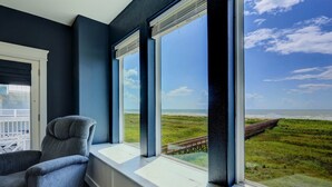 Ein Zimmer mit großem Fenster, Blick auf einen Strand und eine Holzpromenade.