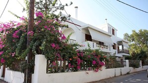 Uma casa branca de dois andares com varanda, cercada por um muro branco e plantas floridas.