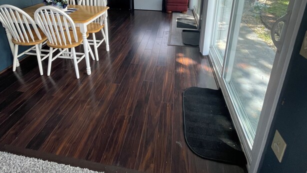 Quarto com mesa de jantar e cadeiras, uma pequena área de estar com um banquinho vermelho, ventoinha no teto e janelas amplas com vista para o exterior.