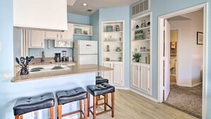 A kitchen with a central island, bar stools, white cabinetry, and a view into the adjacent bathroom. - Corpus Christi Condo - Walk to Whitecap Beach! (Corpus Christi)