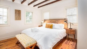 Een slaapkamer met een houten plafond, een groot bed met witte beddengoed, een nachtkastje met een lamp en een bankje.
