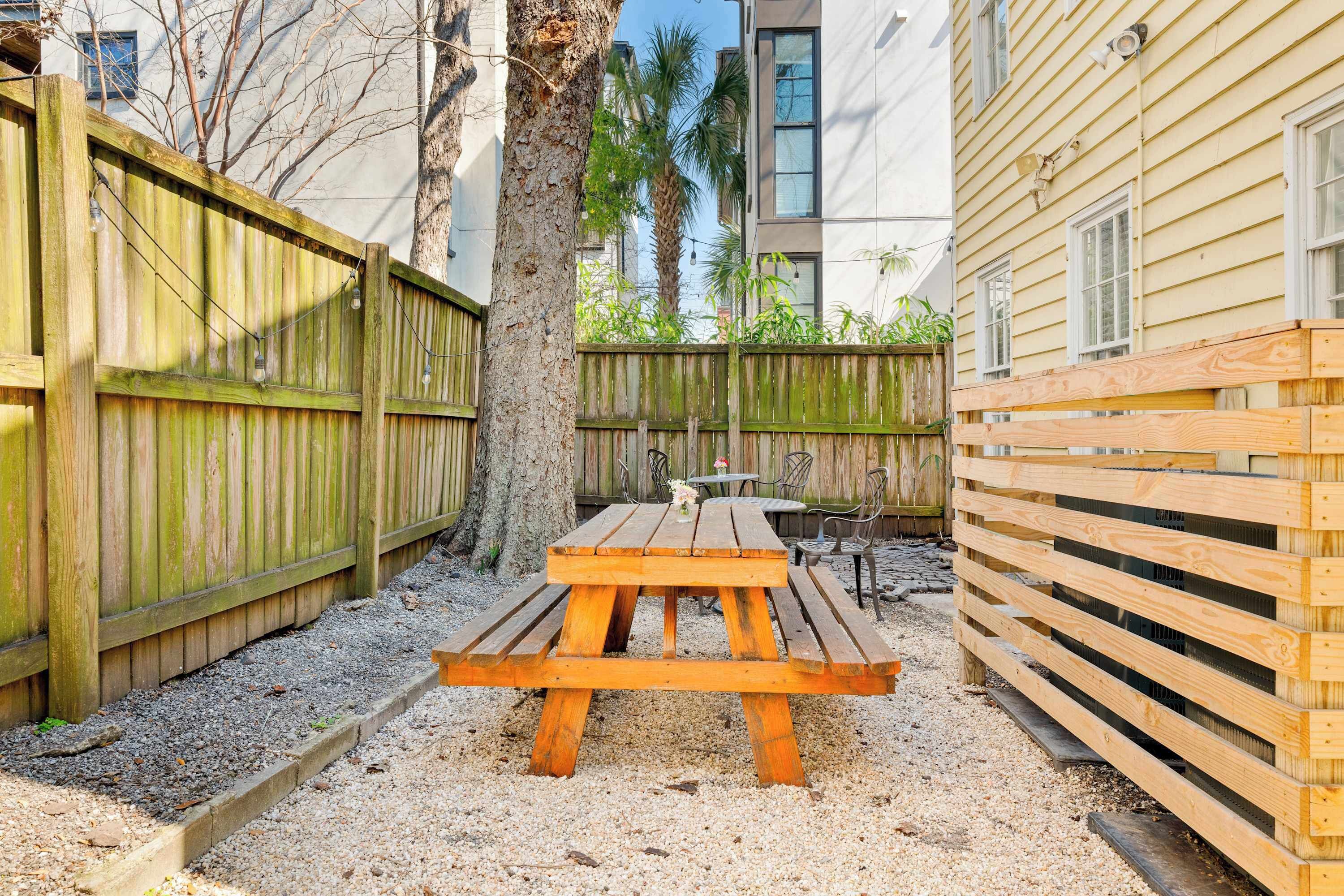 Una mesa de picnic de madera en un patio trasero, con una valla de madera, un árbol y un edificio al fondo.