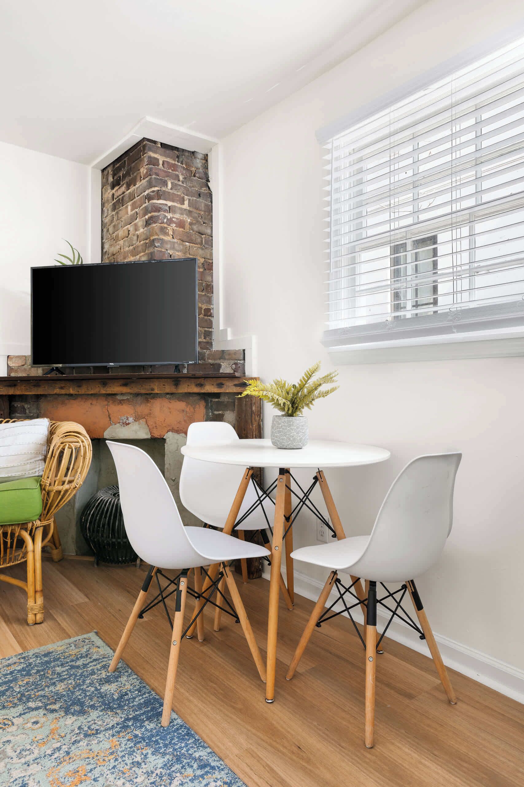 Una sala de estar moderna con chimenea de ladrillo, un televisor de pantalla plana y una zona de comedor con sillas blancas y una mesa pequeña.