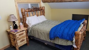 Ein rustikales hölzernes Bett mit einer Decke in Grün- und Blautönen, ein Nachttisch mit Lampe und ein Spiegel.