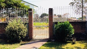 Une entrée fermée par un portail en métal et bordée d’un mur en briques, le tout entouré de verdure et accessible par un chemin pavé.