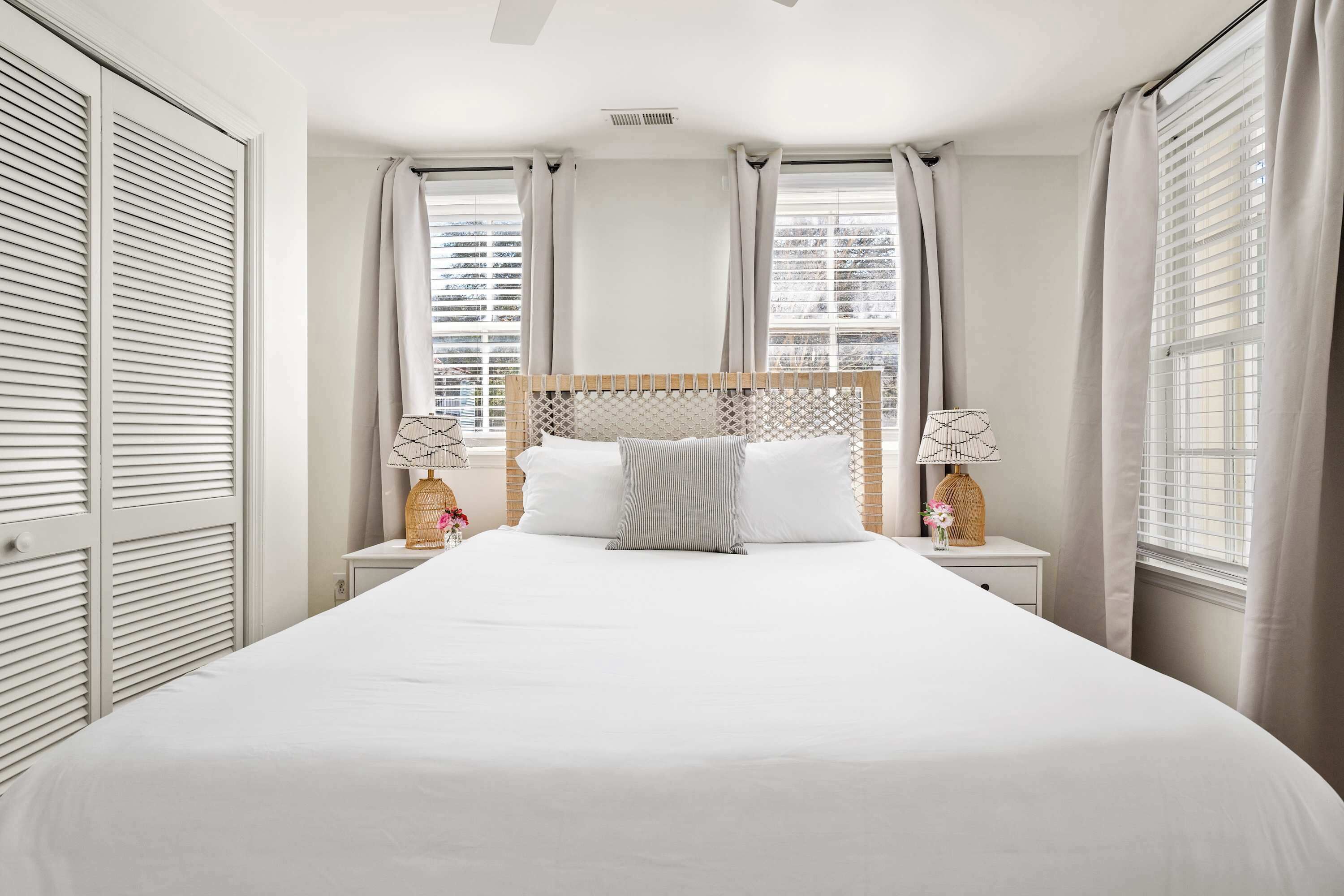 Una cama bien tendida con cabecera de madera, dos mesitas de noche con lámparas y vistas de la ciudad a través de amplias ventanas con cortinas.