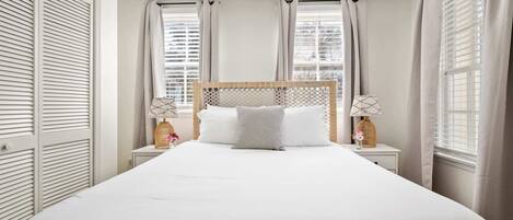 Una cama bien tendida con cabecera de madera, dos mesitas de noche con lámparas y vistas de la ciudad a través de amplias ventanas con cortinas.
