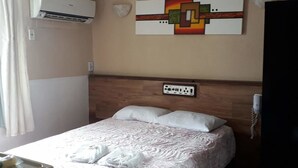 A hotel room with a bed, an air conditioning unit, a power outlet, and a wall-mounted artwork.