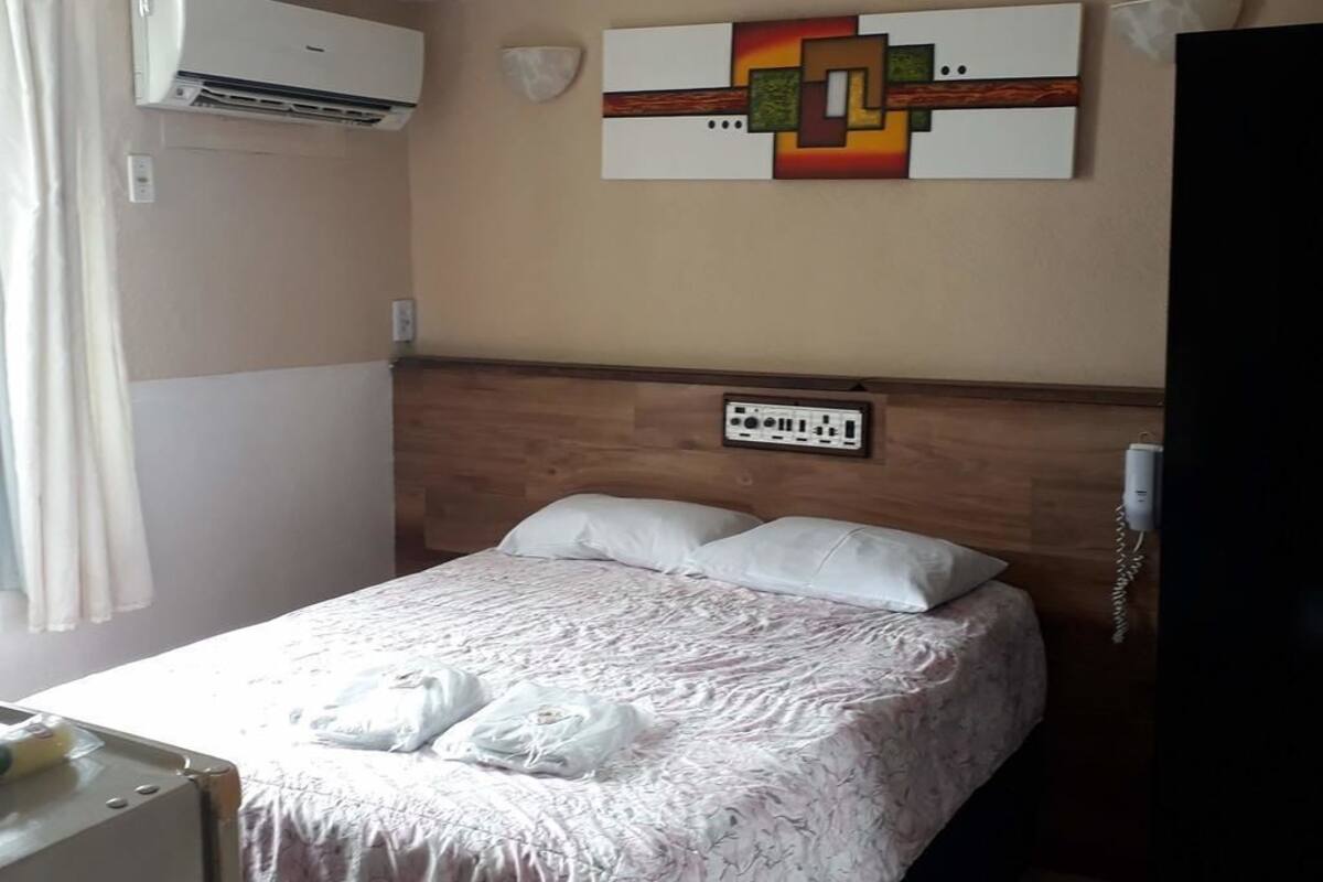 A hotel room with a bed, an air conditioning unit, a power outlet, and a wall-mounted artwork.