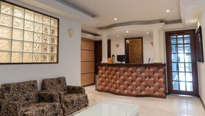 A hotel lobby with a reception desk, patterned sofas, a large decorative window, and a tiled floor.