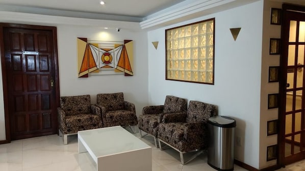 A living room with patterned sofas, a white coffee table, a trash bin, a wooden door, and a wall with abstract art and a window.
