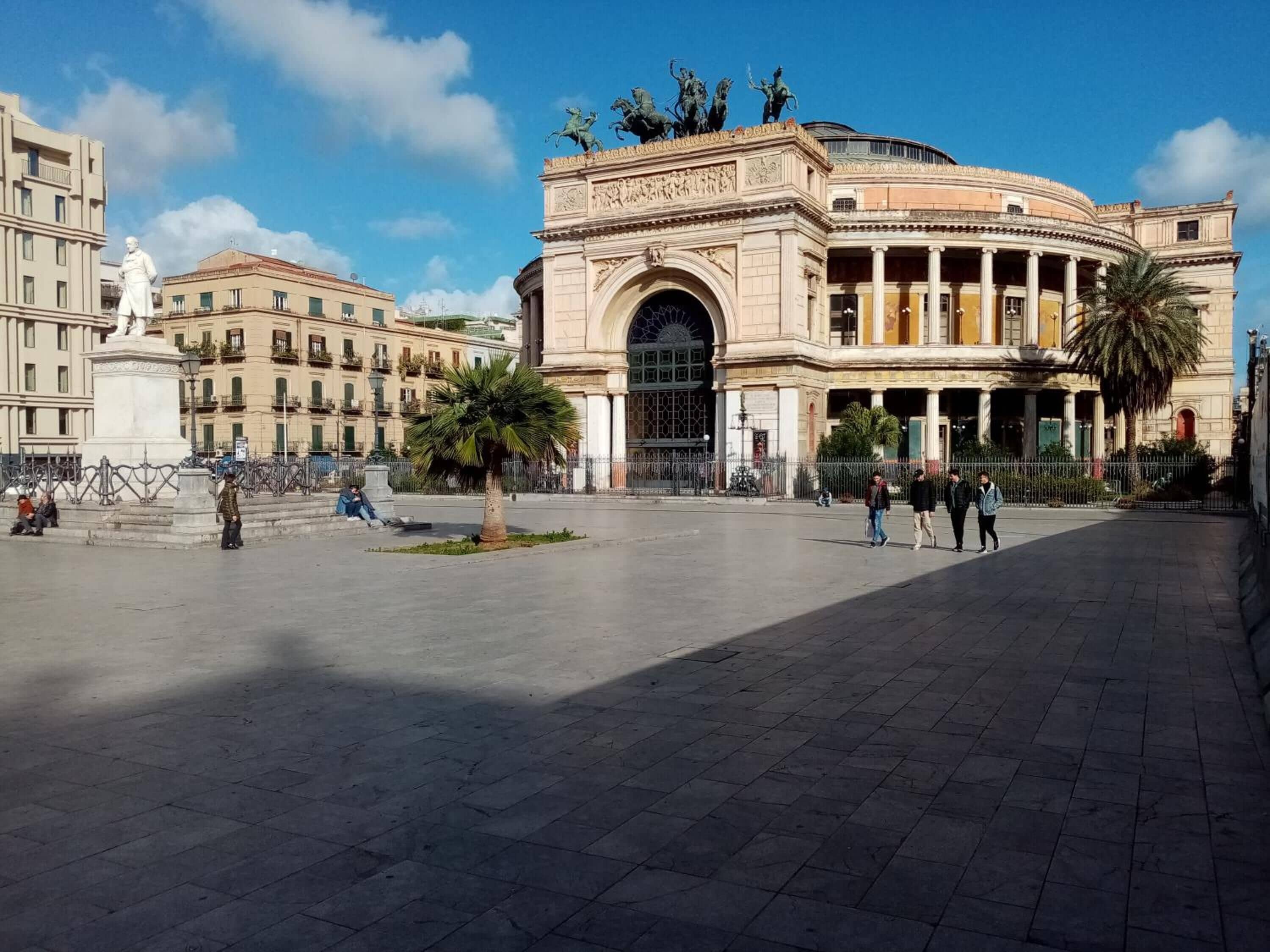 Una plaza pública con un edificio grande de arquitectura clásica, una estatua y palmeras.
