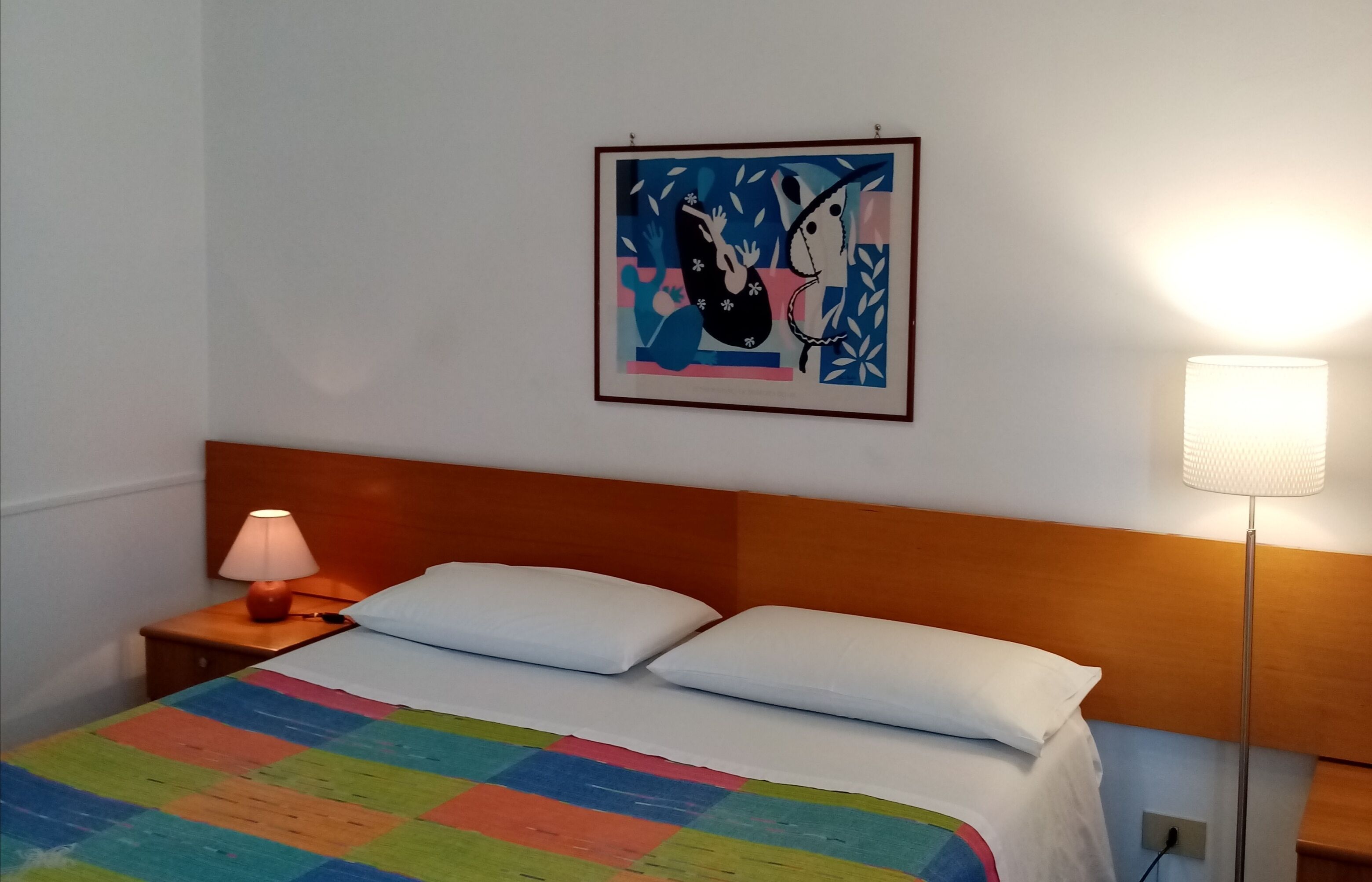 Un lit bien fait, agrémenté d’une couverture colorée, une œuvre d’art encadrée au mur, une lampe de chevet et une tête de lit en bois.