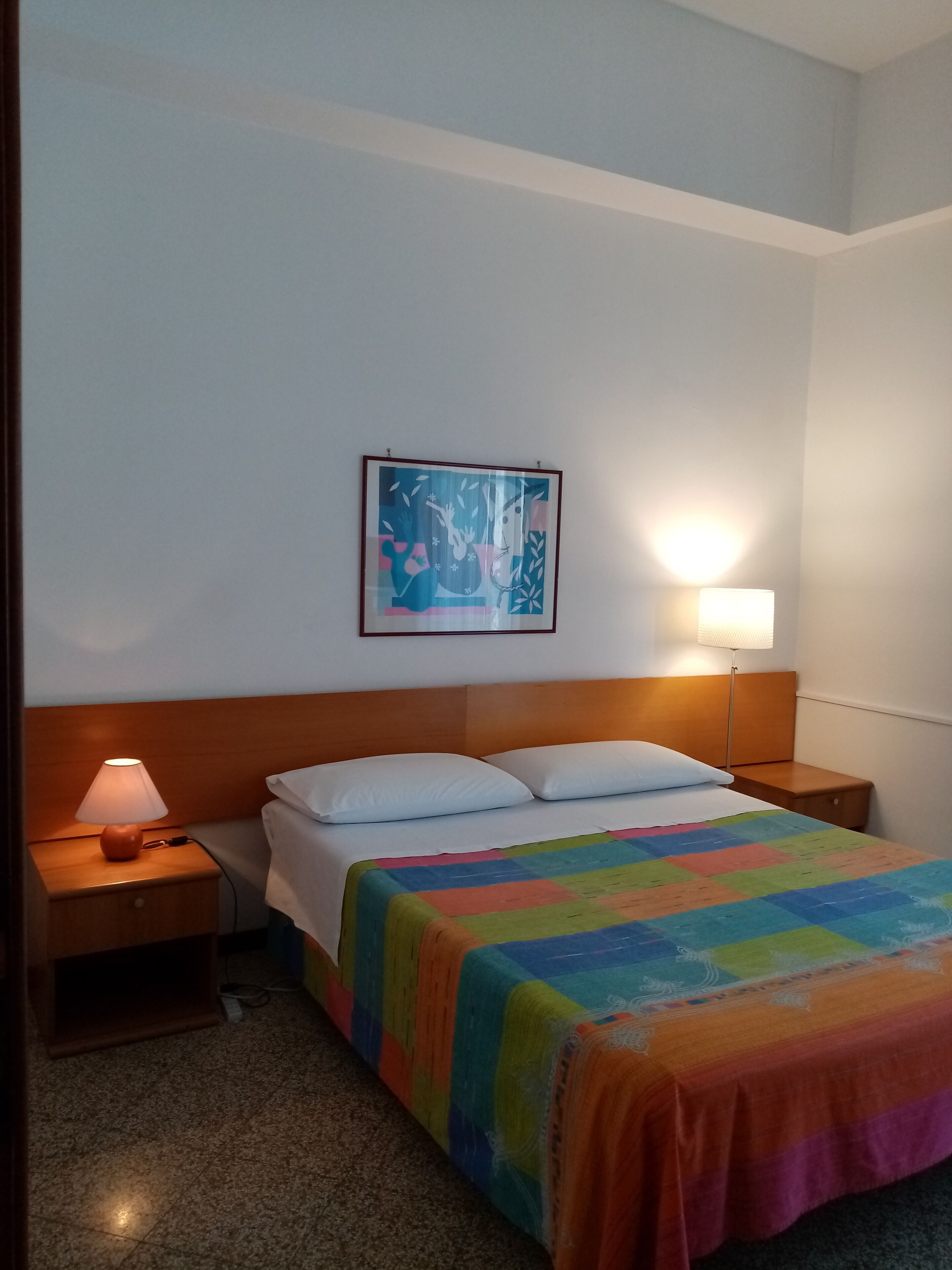 A hotel room with a bed, a nightstand, a lamp, and a wall-mounted painting.
