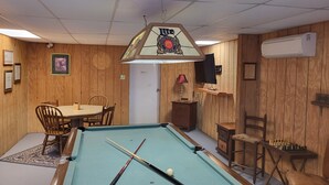 Una mesa de billar con tacos, una mesa con sillas y un televisor en la pared.