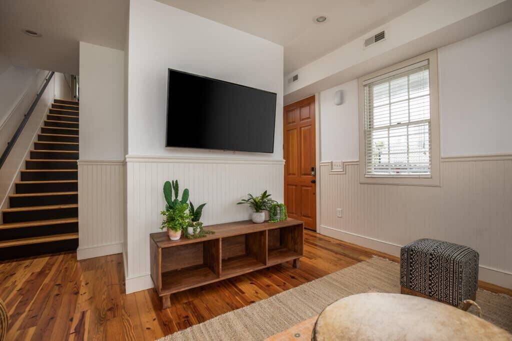 Een woonkamer met een houten vloer, een flatscreen televisie aan de muur, een houten bankje met planten, een gestreept voetenbankje en een trap.
