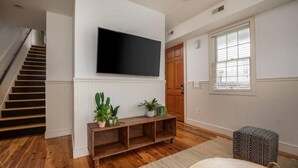 A living room with a wooden floor, a flat-screen TV mounted on the wall, a wooden bench with plants, a patterned ottoman, and a staircase. - 4BR Charleston Single! One Block to King St! (Charleston)