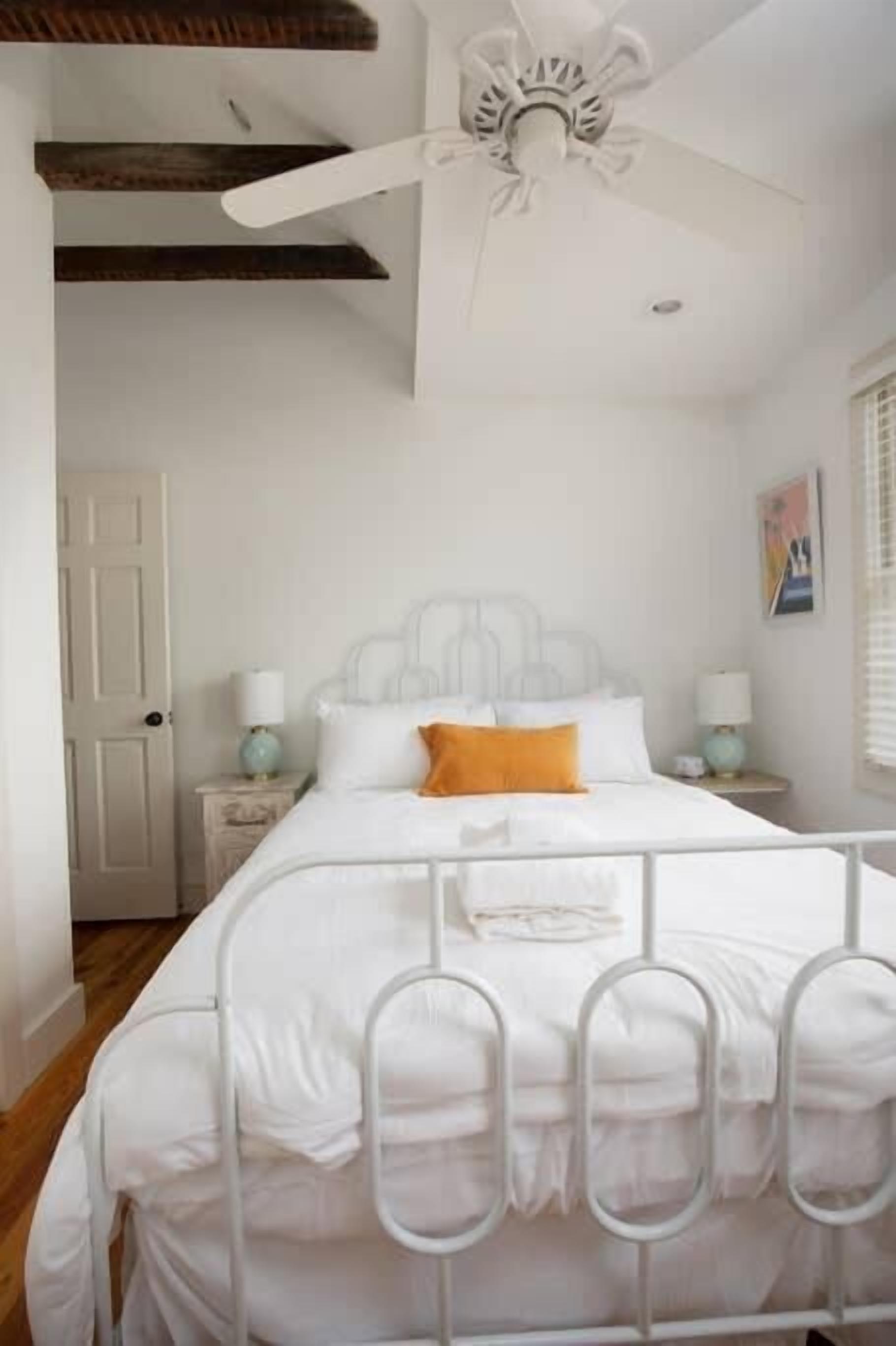 Un dormitorio con una cama blanca, ventilador de techo, suelo de madera y una puerta blanca.