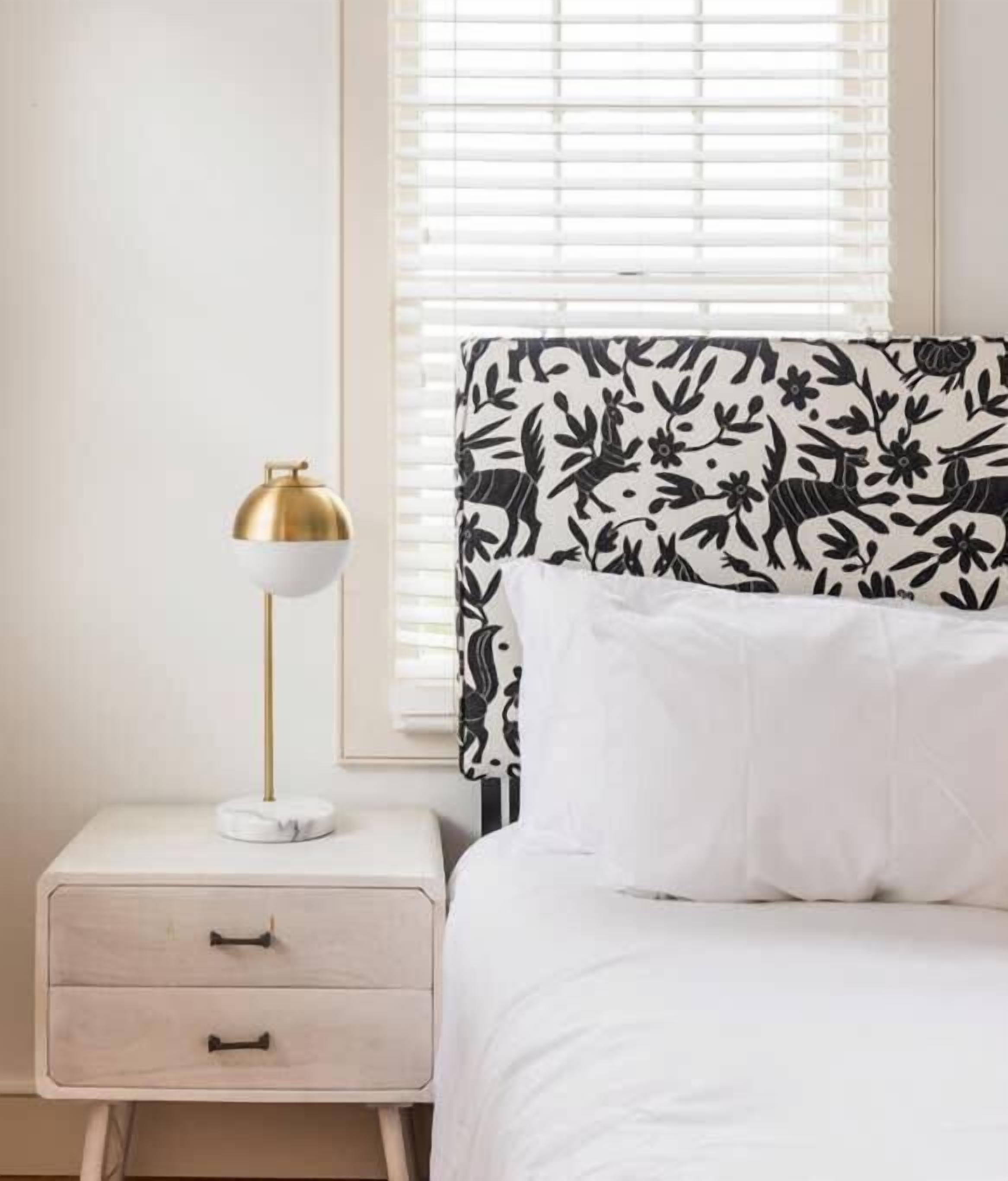 Een slaapkamer met een wit bed, een nachtkastje met lamp en een raam met jaloezieën.