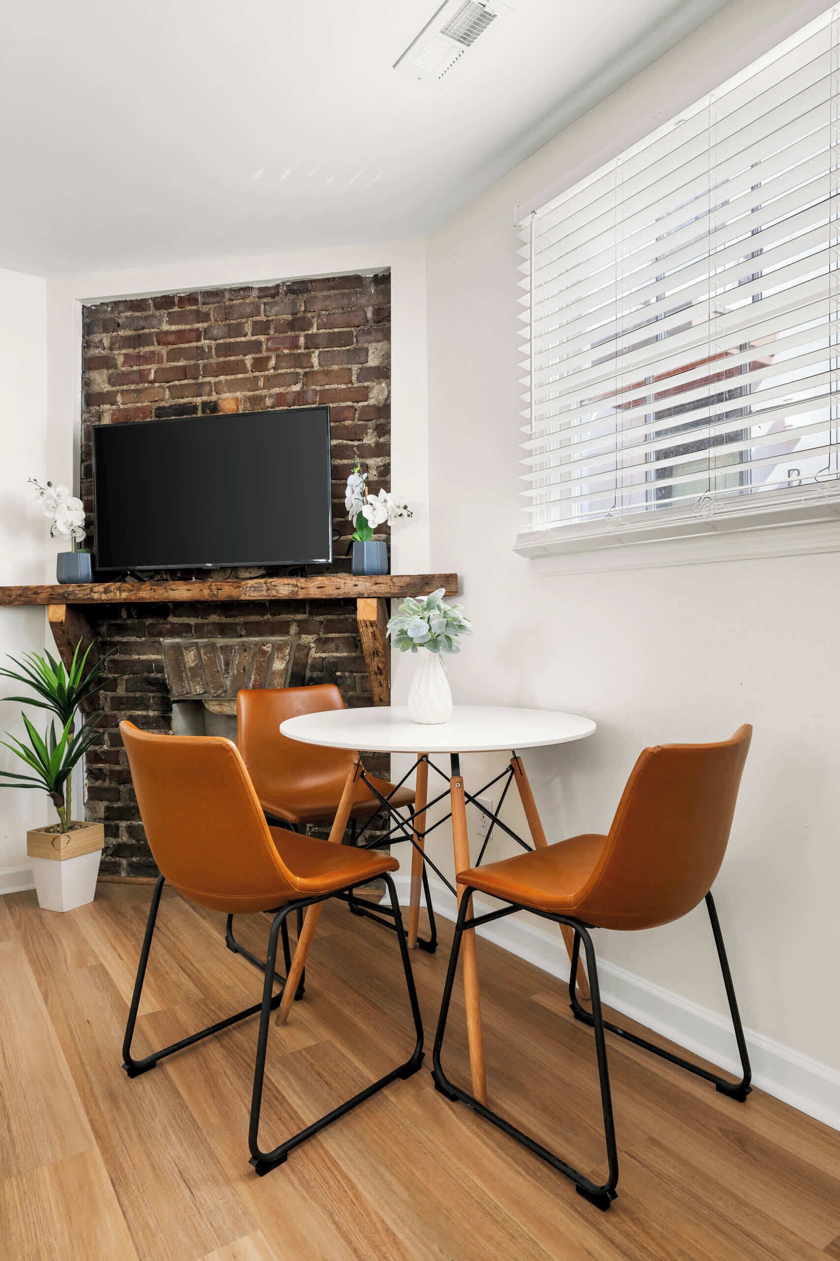 A modern living room with a brick fireplace, a flat-screen TV, and a round dining table with orange chairs.