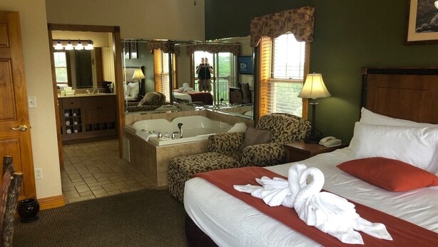 Ein Hotelzimmer mit einem großen Bett, einer Sitzecke mit Sessel und Tisch, einem Badezimmer mit Badewanne und Spiegel sowie einer Tür zu einem weiteren Raum.