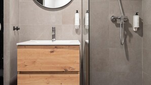 Un baño moderno con un tocador de madera, un espejo redondo, una ducha con cabezal de ducha manual y dos botellas blancas en la pared.