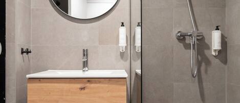 Un baño moderno con un tocador de madera, un espejo redondo, una ducha con cabezal de ducha manual y dos botellas blancas en la pared.