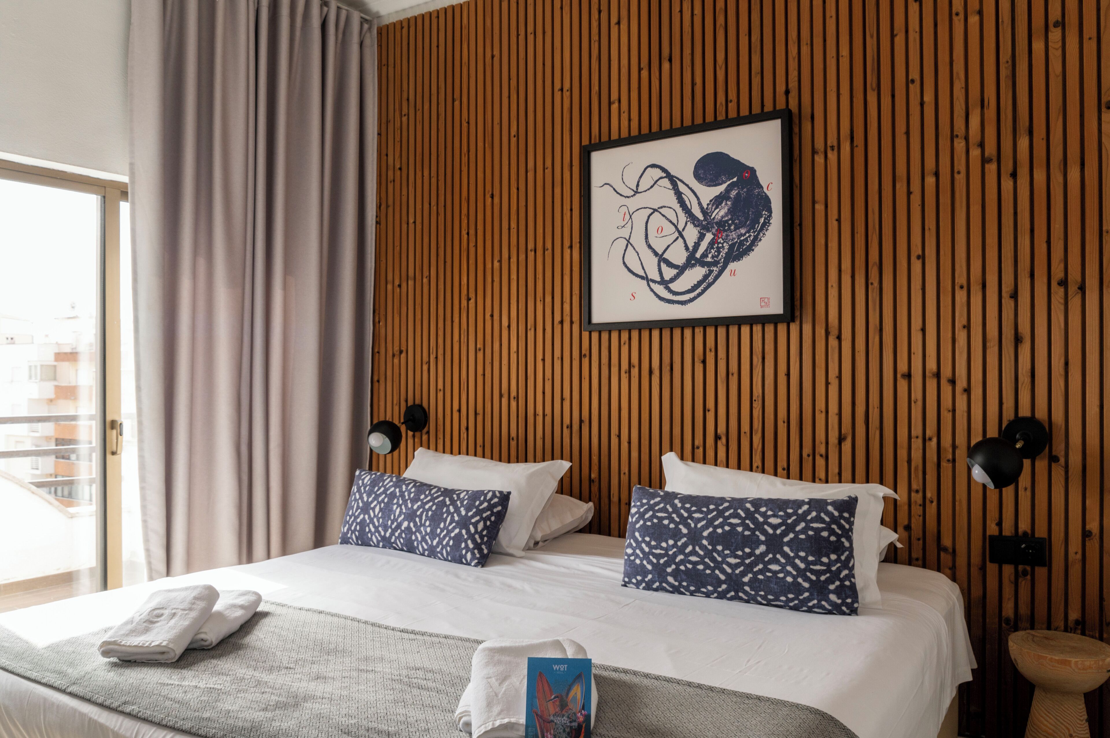 A hotel room with a wooden paneled wall, a bed with white bedding and patterned pillows, a framed artwork, and a bedside lamp.