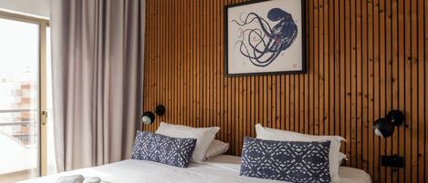 A hotel room with a wooden paneled wall, a bed with white bedding and patterned pillows, a framed artwork, and a bedside lamp.