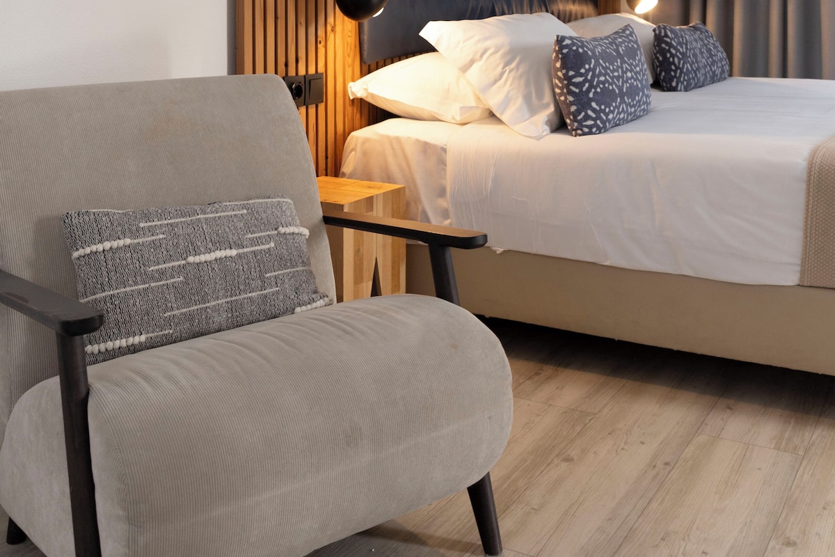 Ein Hotelzimmer mit Bett, Sessel, Schreibtisch, Lampe und zwei gerahmten Bildern an der Wand.
