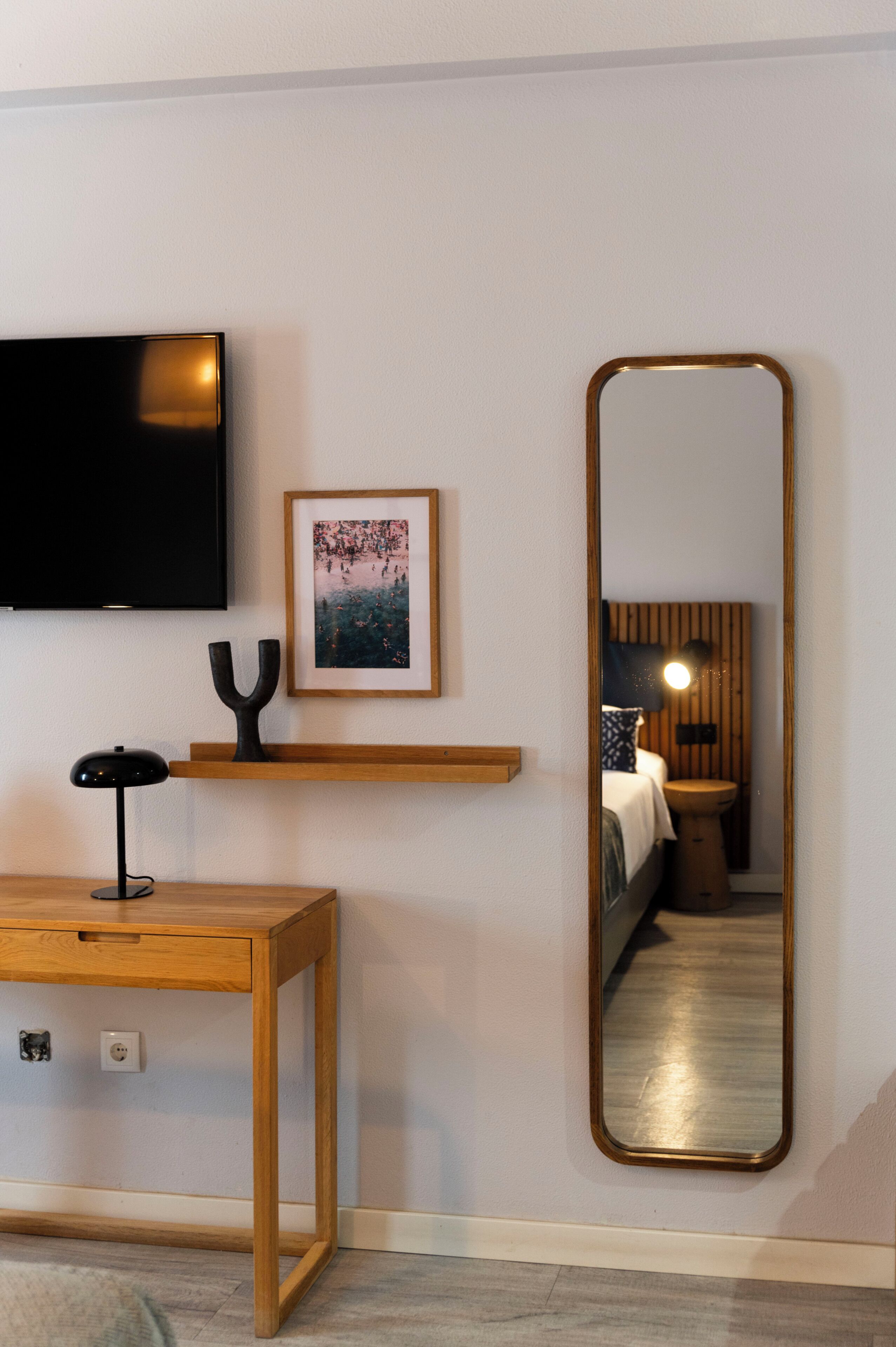 Una habitación con televisión montada en la pared, un cuadro enmarco, un escritorio de madera con una lámpara y un espejo grande que refleja una cama y una mesita de noche.