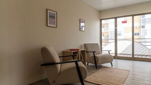Een moderne hotelkamer met twee beige fauteuils, een klein houten bijzettafeltje en een groot raam met uitzicht op de buitenkant van het gebouw.