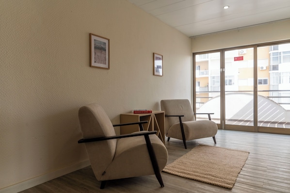 Una habitación de hotel moderna con dos sillones color beige, una pequeña mesita de madera y un ventanal que ofrece vistas del exterior del edificio.