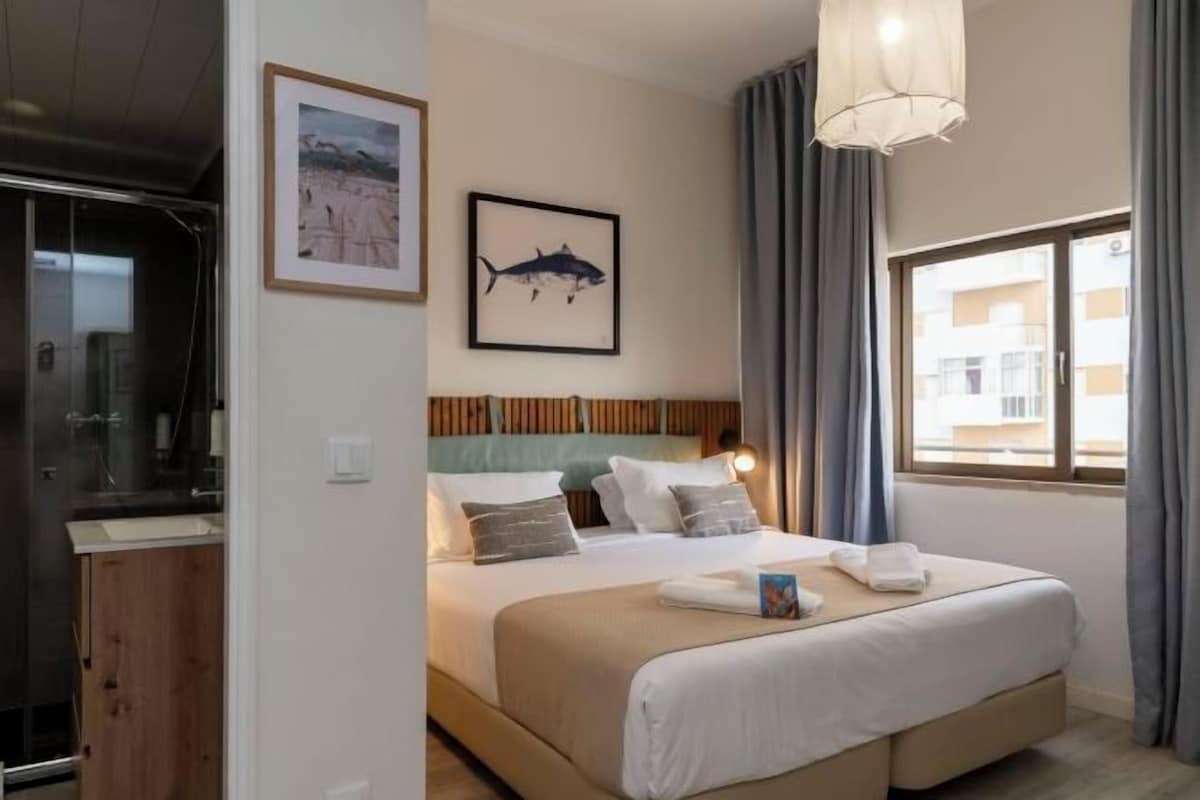 Ein Hotelzimmer mit Bett, Kopfteil, einem Fenster mit Vorhängen, einem gerahmten Bild an der Wand und einem Badezimmer mit Dusche.